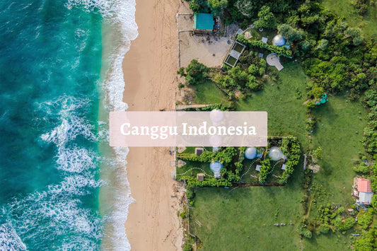 Aerial view of Canggu Indonesia's turquoise surf rolling onto a sandy beach beside lush green fields and seaside huts, capturing Bali’s laid-back coastal vibe.